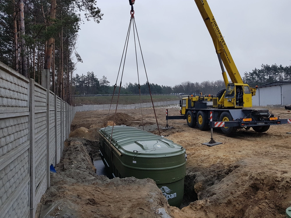 Żółty dźwig opuszcza zielony zbiornik przydomowej oczyszczalni ścieków do wykopu na placu budowy