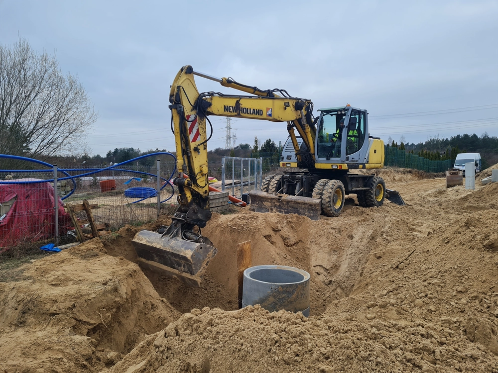 Żółta koparka wykonuje prace ziemne przy wykopie i montażu betonowego pierścienia studni