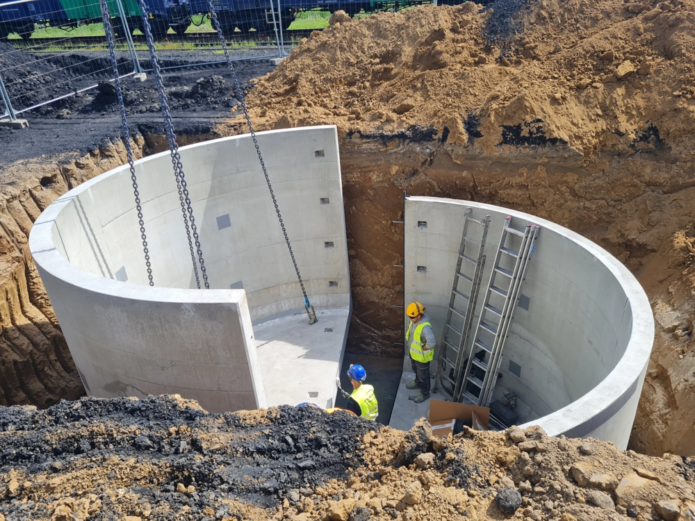 Montaż prefabrykowanej przepompowni ścieków w wykopie, betonowe elementy i pracownicy przy pracach ziemnych