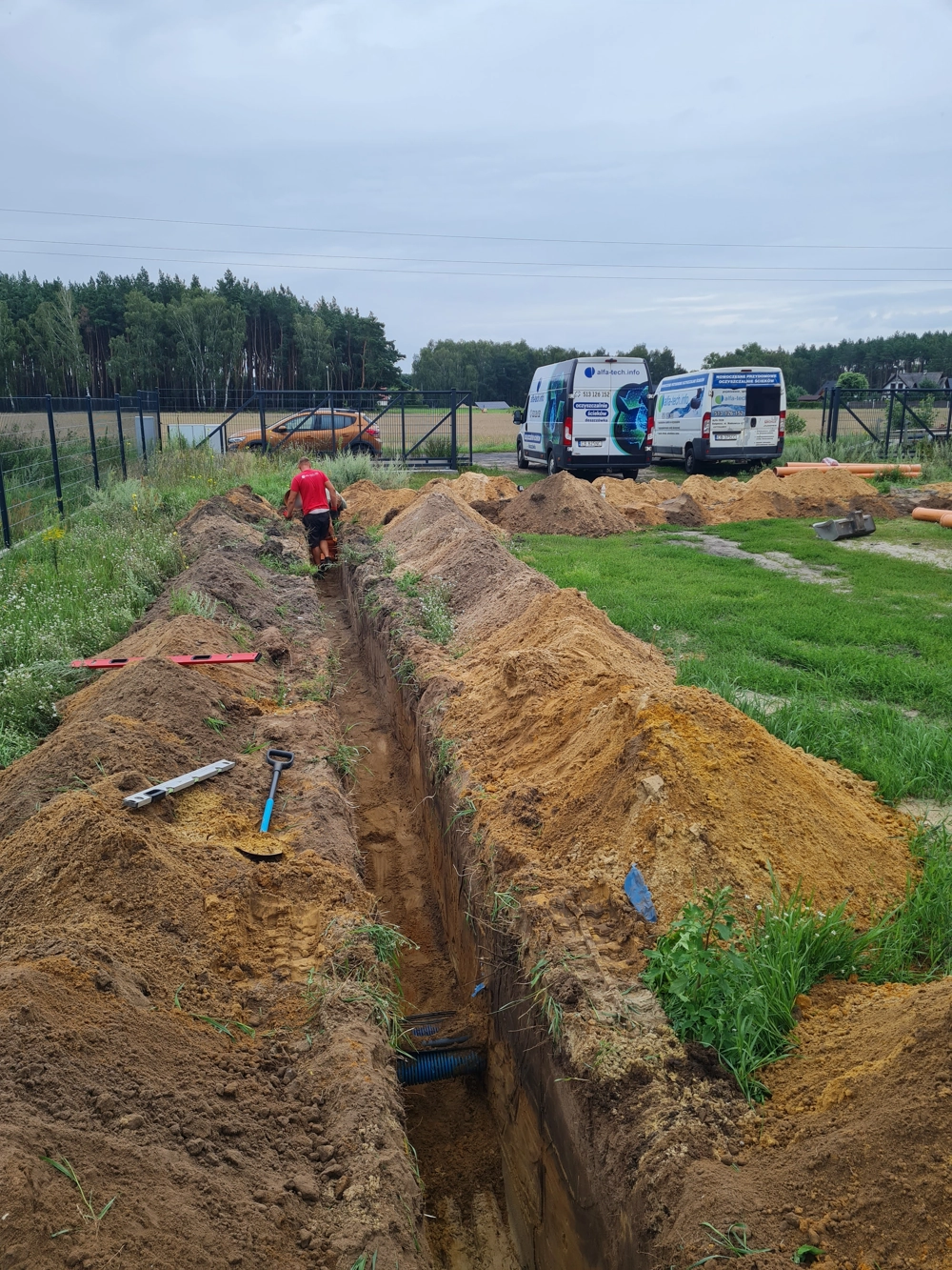 Wykop pod drenaż – układanie rury odwadniającej w wykopie, widoczne narzędzia i vany serwisowe