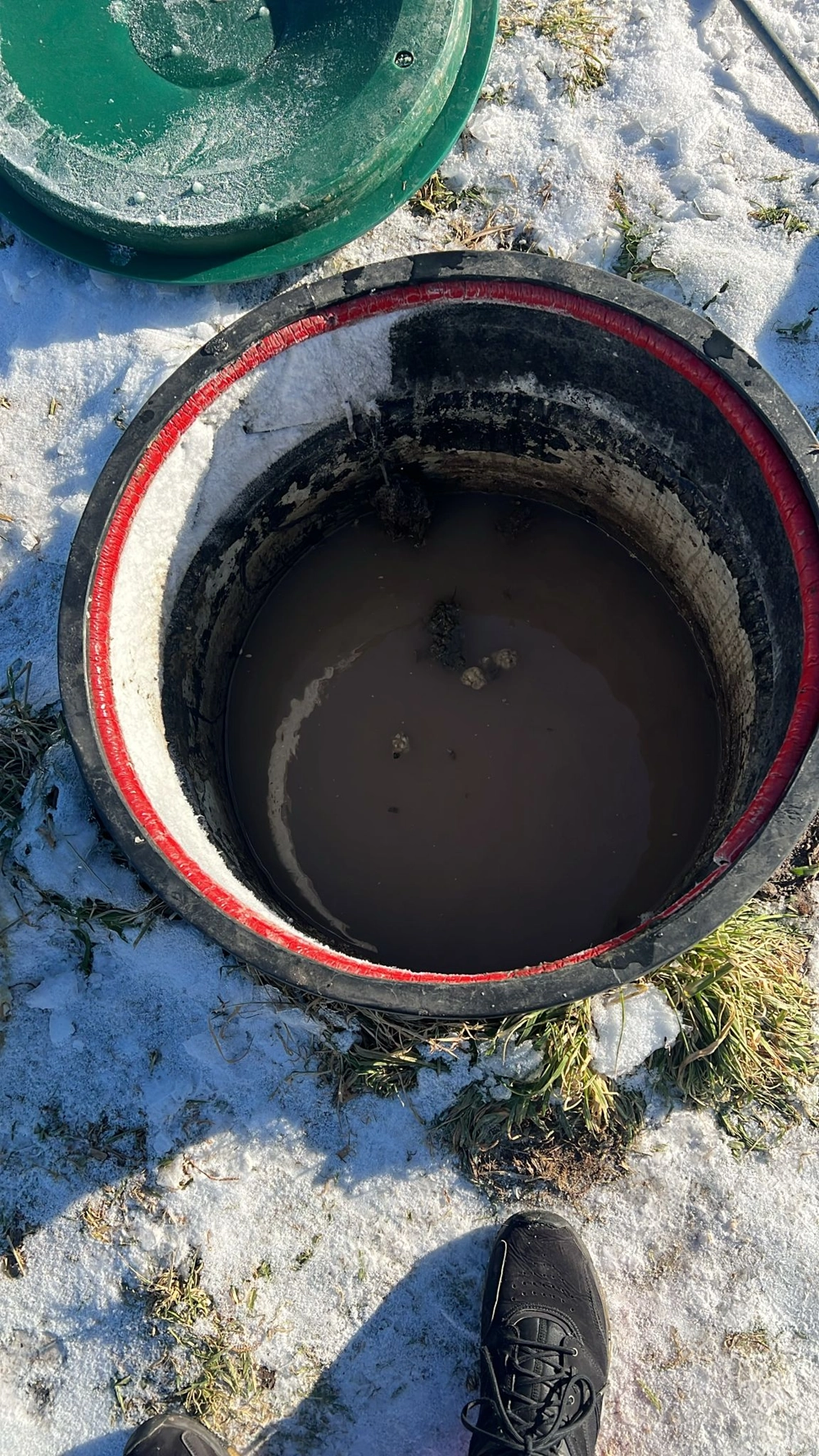 Otwarty właz szamba betonowego z brunatną wodą, na zaśnieżonym terenie podczas prac ziemnych