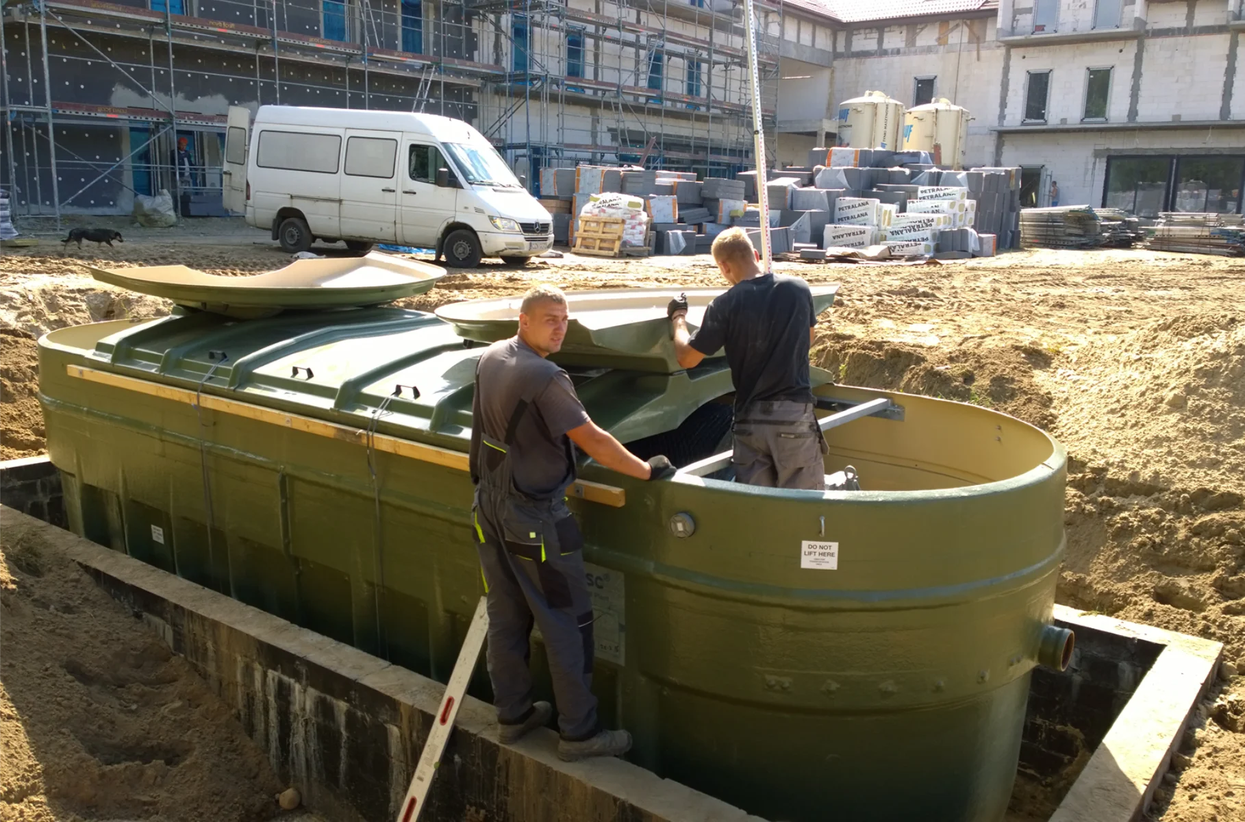 Dwóch pracowników montuje przydomową oczyszczalnię ścieków w wykopie na placu budowy
