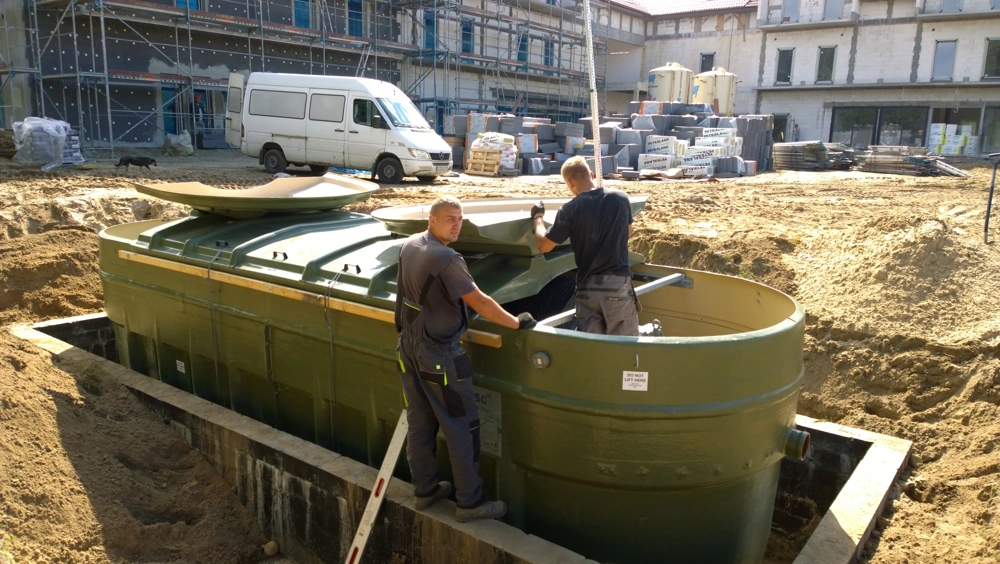 Instalacja przydomowej oczyszczalni ścieków w wykopie - montaż zielonego zbiornika przez dwóch pracowników na placu budowy